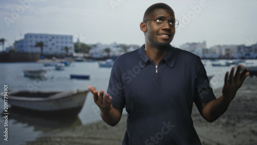 Cuadro en lienzo Man with glasses gestures open hands toward small boats at a coastal harbor beside a white building, smiling in daylight; welcoming warmth