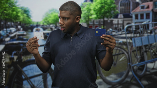 Принти на полотні Man holding two creditcard, comparing a blue card and a white chip card with a puzzled look on a canal street; doubt decision