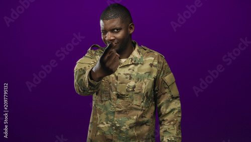 Фотография Soldier man in camouflage uniform beckons with fingers toward camera, prominent hand visible against purple studio wall; pride duty