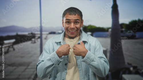 Konstfotografi Man smiling and thumbs pointing to chest in denim jacket on a seaside street with benches and palm trees; playful confidence