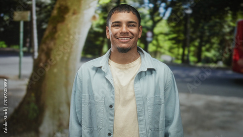 Fotografía Man smiling with visible face in a light blue denim shirt standing beside a large tree on a street near a red bus and sidewalk; warm optimism