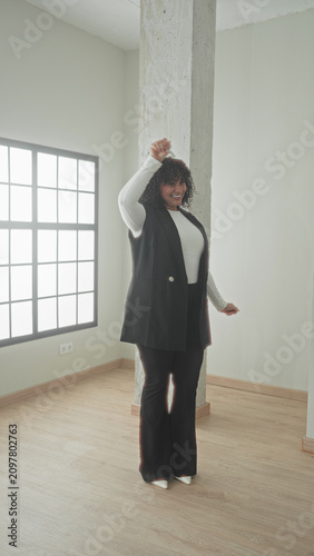 Canvas Print Woman holding key aloft and smiling with raised arm in bright modern building empty room; joy new home success