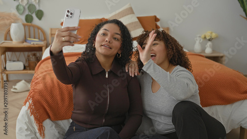Photos Woman holds smartphone aloft taking a selfie while companion rests her head on the other's shoulder in a cozy bedroom with orange bedding; joy friendship