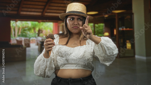 Photos Plus size woman holding chocolate ice cream cone, pointing finger forward, exposed stomach visible, wearing straw hat and white blouse in building lobby; annoyance refusal