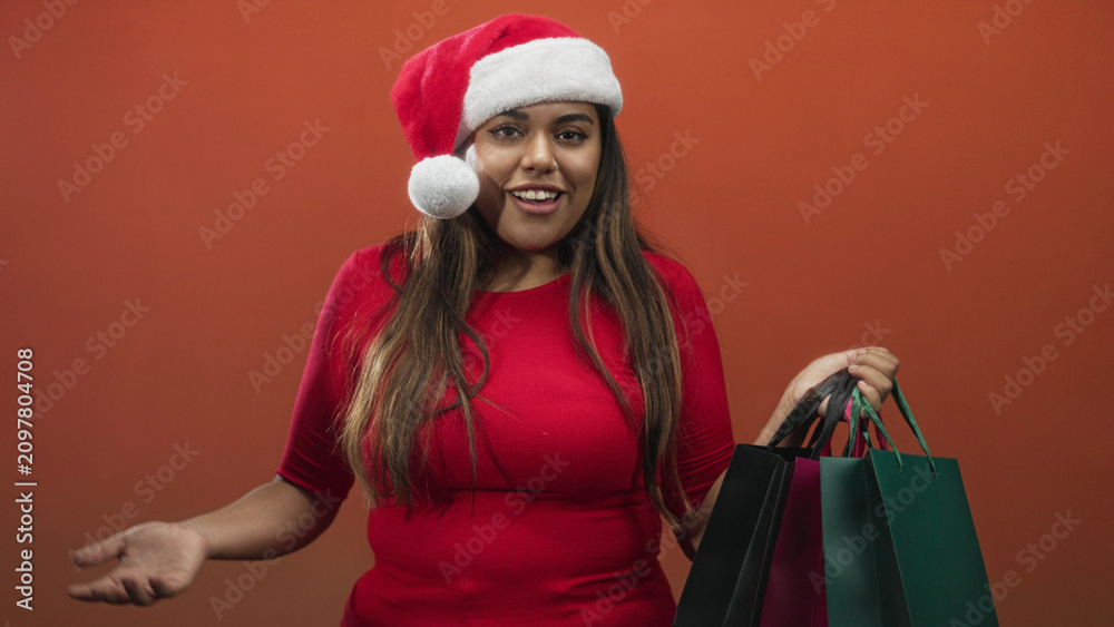 Fototapeta premium Woman wearing santa hat holds multiple shopping bags aloft with raised hand and open mouth expression in studio; holiday shopping joy.