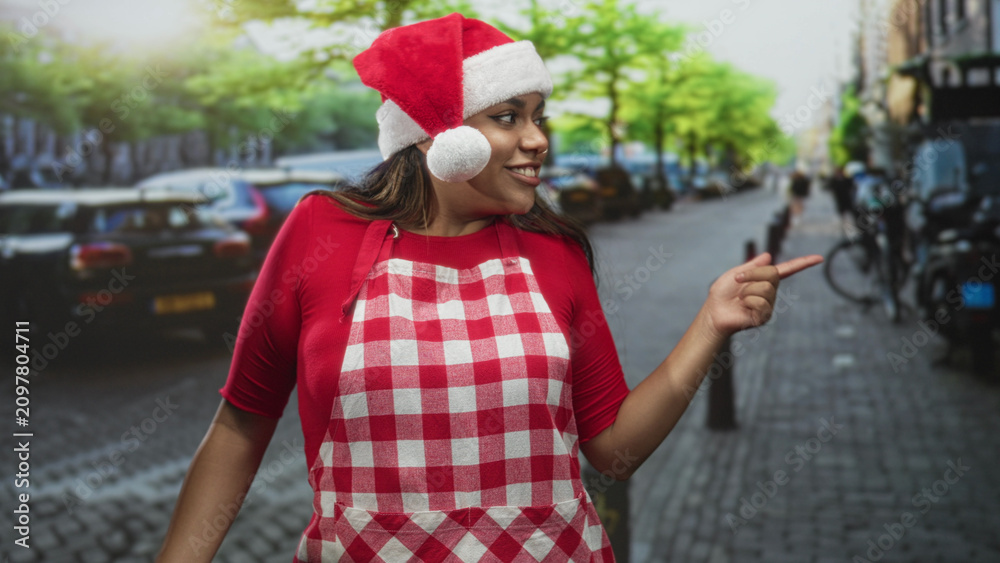 Fototapeta premium Woman in santa hat and red checked apron points finger on street while smiling with closed eyes; festive joy.