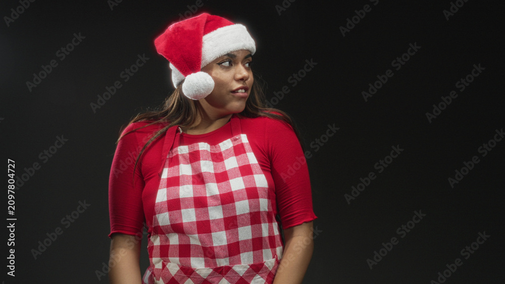 Fototapeta premium Woman in santa hat and red check apron, eyes closed and hands clasped in studio; holiday cheer contentment.