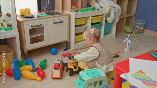 Photography Little blonde girl plays with toy cars, holding a green plastic screwdriver and arranging blocks on a red table in a preschool playroom; focused curiosity