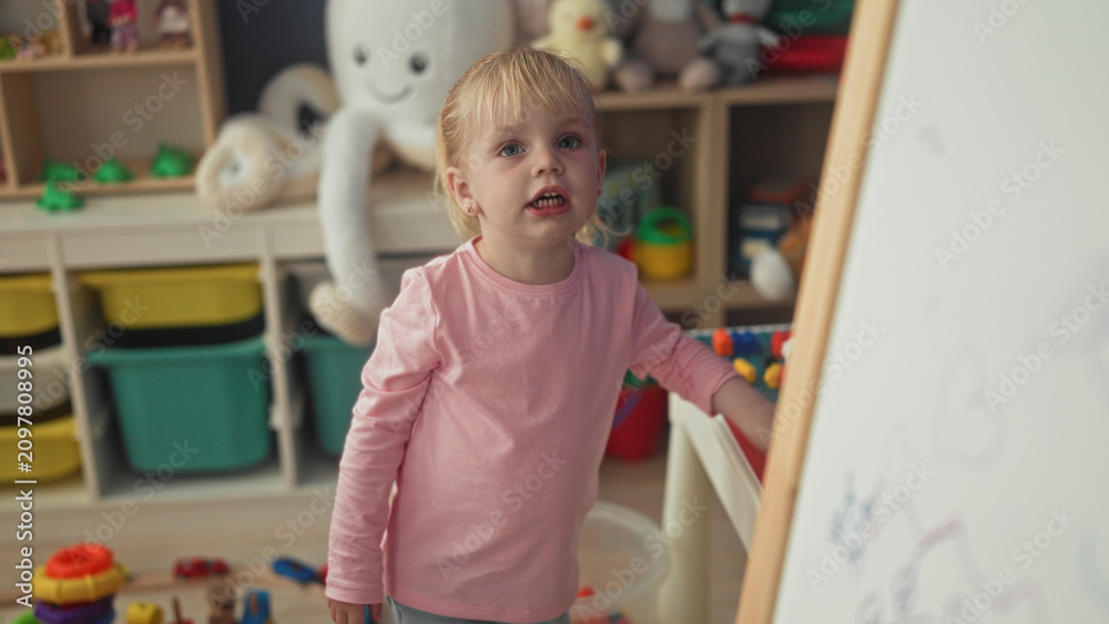Obraz premium Little blonde girl reaches into toy bin and touches easel in a playroom building; playful curiosity.