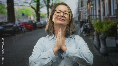 Foto Woman senior with grey hair and glasses pressing palms together in prayer on a cobblestone street beside parked bikes and cars, wearing light blue shirt; gratitude hope