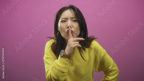 Photos Woman in yellow sweater holds finger to lips shushing in studio against pink wall; secretive hush silence
