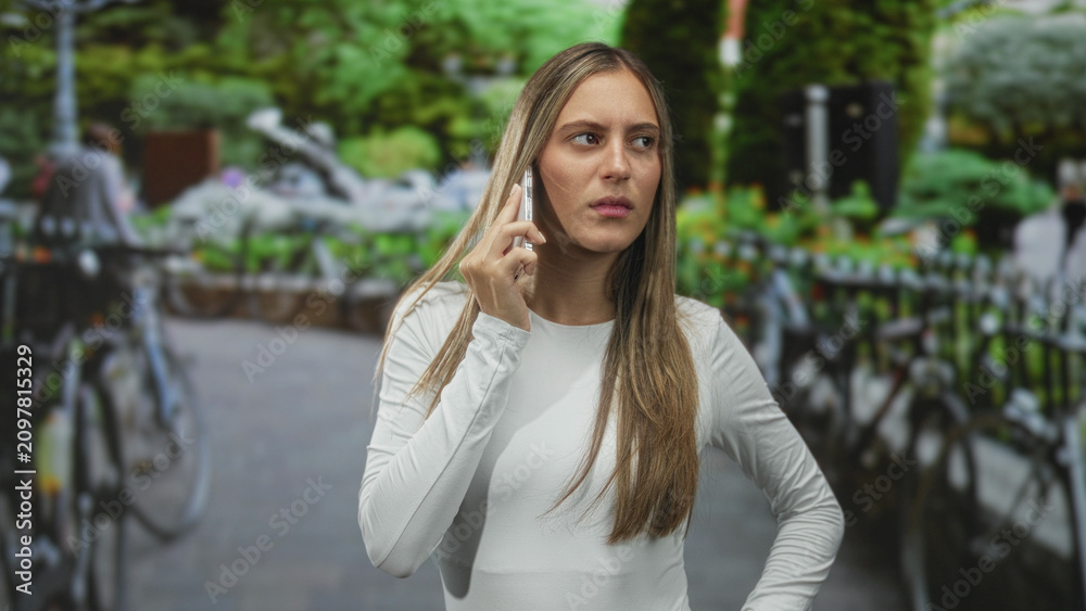 Fototapeta premium Woman holding smartphone to ear, rolling eyes and frowning while standing on a city street beside parked bicycles; annoyance phone call.