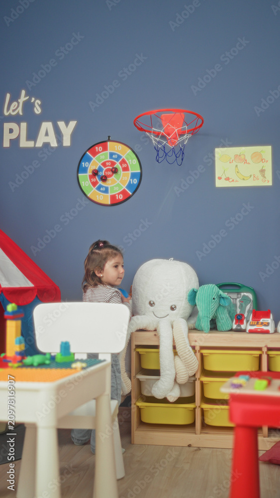 Obraz premium Girl holds ball and peeks behind toy shelf in colorful playroom with stuffed octopus, green elephant plush, block table and wall hoop; playful joy.