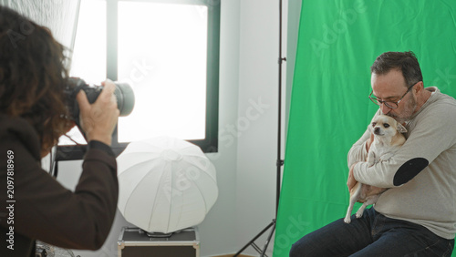 Quadro su tela Man cradling small chihuahua on stool, petting its head while a photographer shoots near a lighting umbrella in studio with green backdrop; warm affection