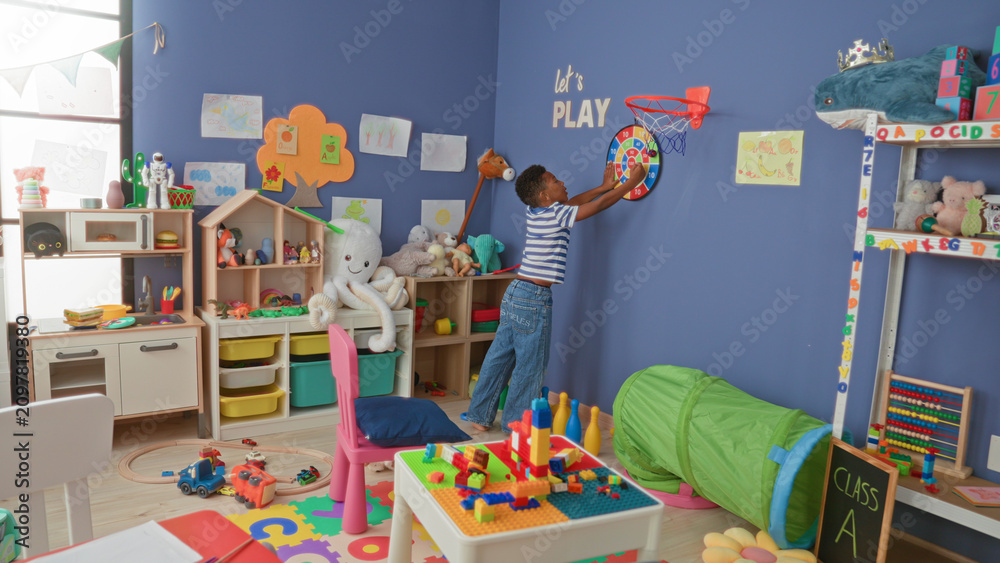 Fototapeta premium African american boy reaching for wall dartboard while holding toy car in colorful classroom; playful curiosity.
