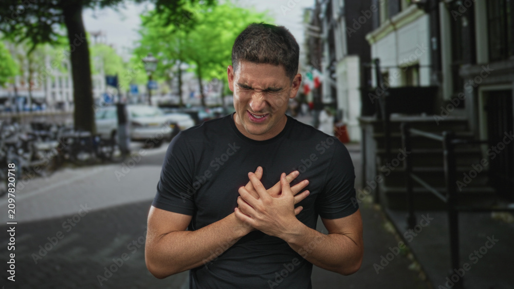 Fototapeta premium Young hispanic man grimacing clutching chest with bare hands on city street near bicycles and parked cars; heartattack emergency pain.