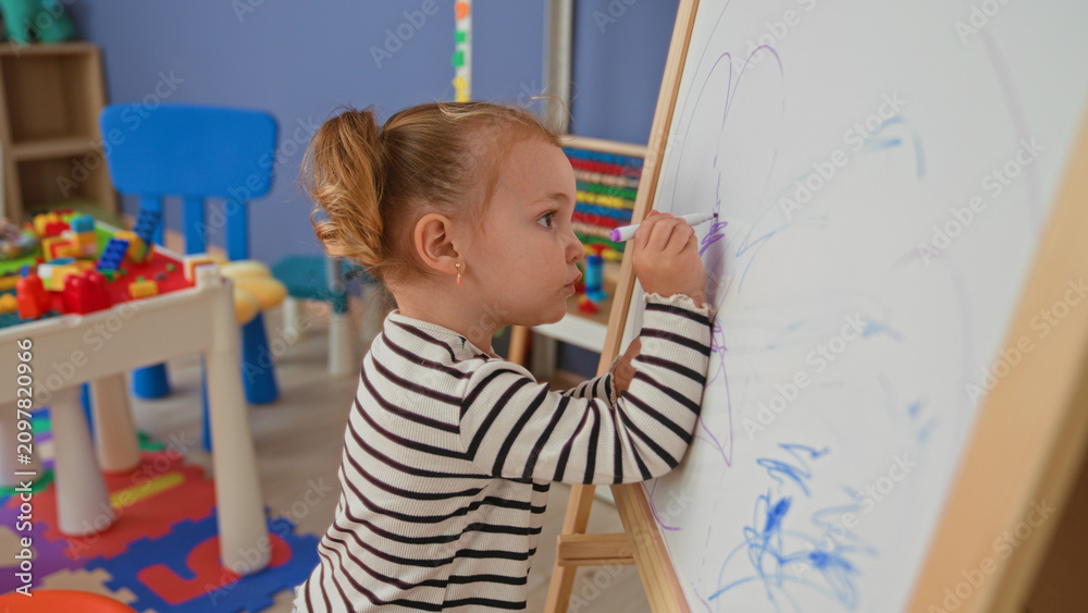 Fototapeta premium Little blonde girl drawing on a whiteboard with a purple marker at a wooden easel in a daycare building, small hand on board and intent expression; curiosity learning play.