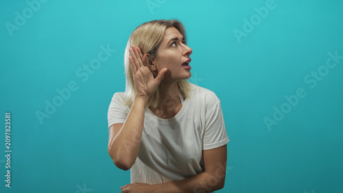 Quadro su tela Young blonde woman cupping hand to ear in studio, wearing white tshirt and gazing sideways with open mouth listening gesture; curiosity