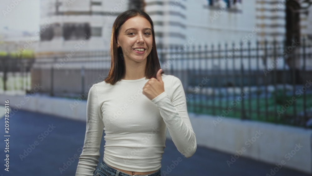 Fototapeta premium Young hispanic woman smiling, hand raised showing thumbs up and exposed midriff on a city street in front of an iron fence and stone building; friendly confidence.
