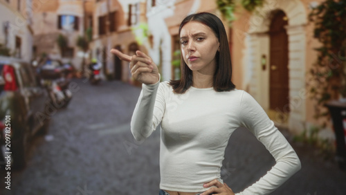 Obraz na plátně Young woman points finger with hand on hip in cobblestone street, skeptical expression and furrowed brow; skepticism