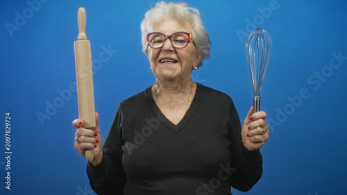 Fotografie Grandmother holding a wooden rolling pin and metal whisk with red nails, smiling and gesturing with hands in a studio portrait; nostalgia joy