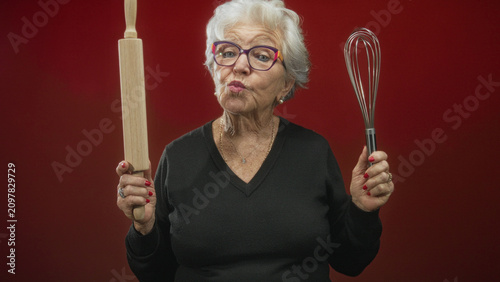 Bild auf Leinwand Elderly woman wearing colorful glasses holds a whisk and rolling pin in hands, pouts lips toward camera in studio; playful confidence
