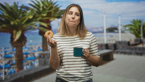Konstfotografi Woman holding a glazed donut and blue mug, tongue out and smiling in a striped shirt on a street boardwalk by palm trees and beach umbrellas; playful relaxation