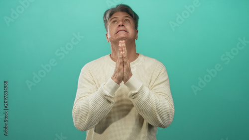 Schilderij op canvas Senior man with hands pressed together in prayer gesture in studio teal backdrop, eyes closed and head bowed; quiet contemplation serenity