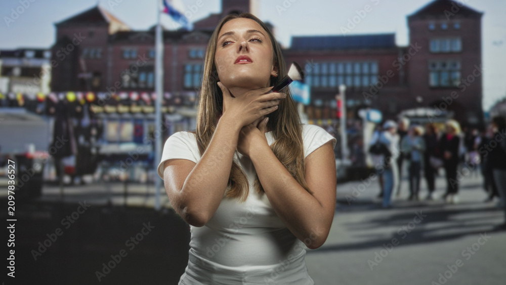 Fototapeta premium Young brunette woman holding a makeup brush to her neck and smiling on a busy street with pedestrians and vendor stalls nearby; quiet confidence.