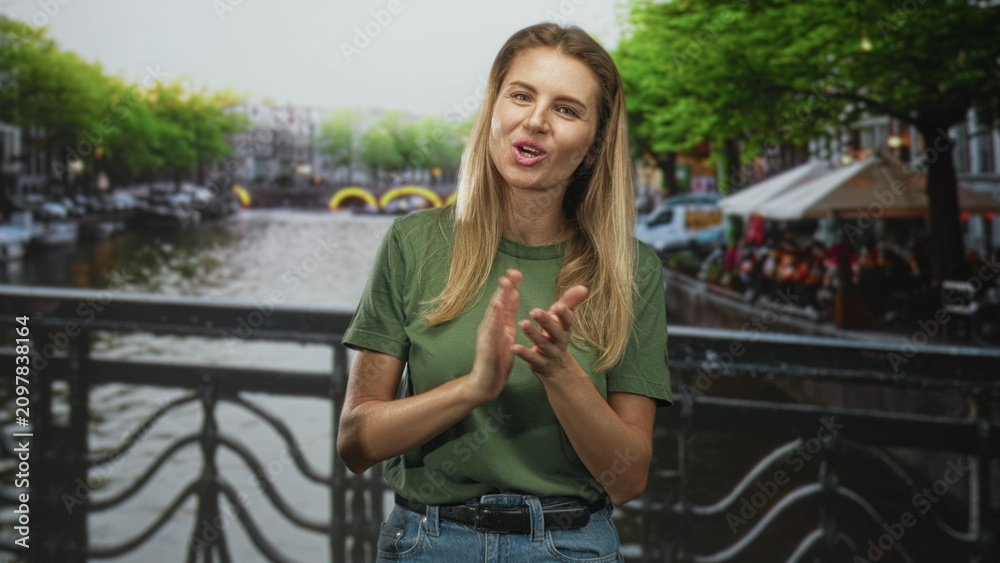 Fototapeta premium Woman clapping hands on a canal bridge by a street cafe with trees and water in view; celebration joy.