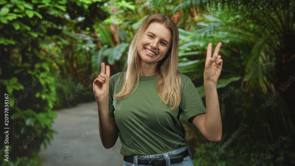 Fototapeta premium Blonde woman holds up victory signs with both hands on a forest path wearing a green t shirt and jeans; joy peace freedom.