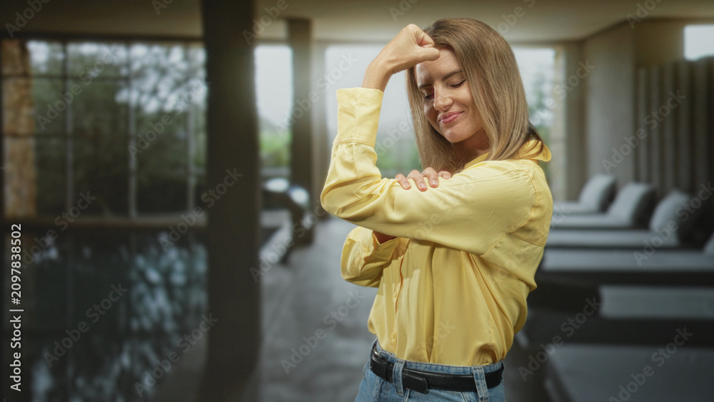 Fototapeta premium Woman flexes bicep with folded arm in yellow shirt at an indoor spa pool lounge inside a building; confidence empowerment.