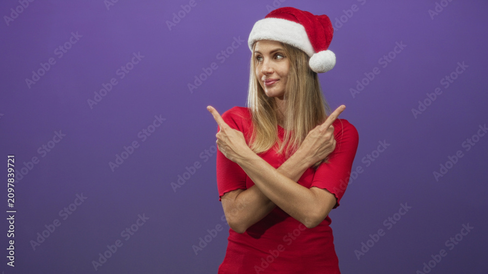 Fototapeta premium Woman in santa hat points crossed fingers while arms are crossed in studio against purple backdrop wearing red top; playful holiday cheer.