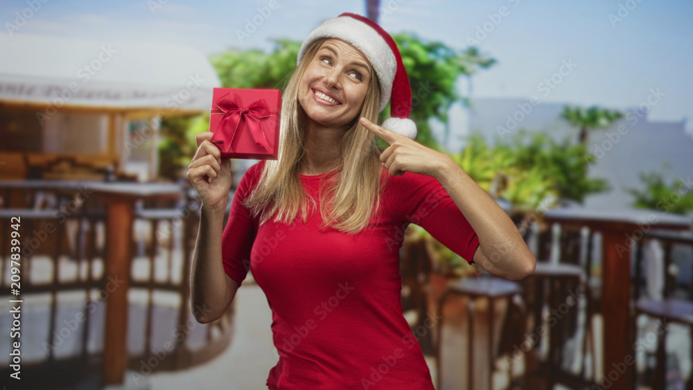 Fototapeta premium Woman in santa hat and red top holds a wrapped red gift with ribbon, smiles and points finger to the gift against a patio backdrop in a studio; festive joy.