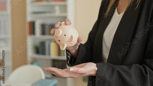 Canvas Print Woman holding pale piggy bank above open palm inside building, wearing business blazer and ring, gesturing offering savings; financial prudence