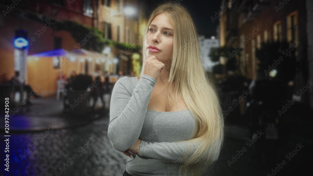 Fototapeta premium Young blonde woman with hand on chin and folded arm wearing a grey long sleeve top, standing by a cobblestone street cafe under lights at night; contemplation.
