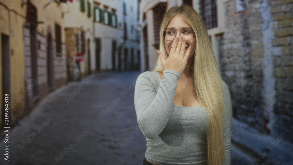 Fototapeta premium Young blonde woman with glasses covers mouth with hand on narrow cobblestone street; surprise reaction.