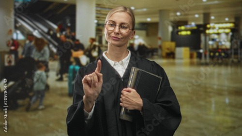 Tablou pe pânză Middle aged blonde woman judge in robe points finger while holding law book in busy airport terminal; authority resolve