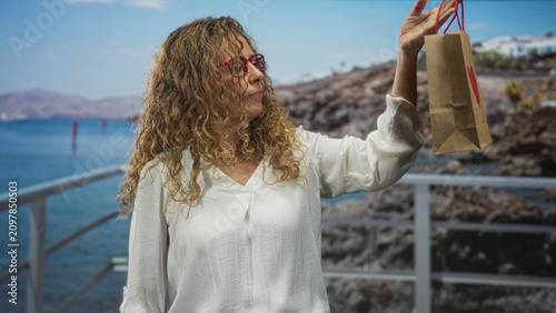 Cuadro en lienzo Woman in white blouse and red glasses holds small brown paper gift bag with heart aloft by right hand on seaside pier railing; gentle nostalgia