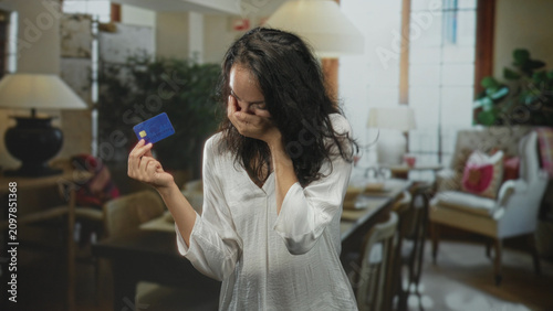 Obraz na plátne Woman holding blue creditcard and hand covering face in a building living room, expressing distress over finances; financial anxiety