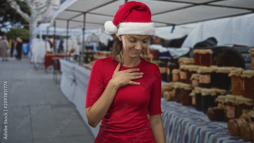 Fototapeta premium Young hispanic woman in santa hat touches hand to chest at a street market stall with jars and canopy; holiday gratitude.