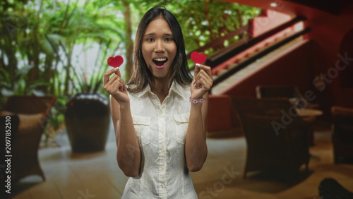 Foto Woman holding two red heart props in each hand inside a lobby building with worried, tense expression and pleading gesture; romantic anxiety