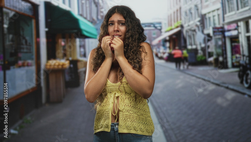 Foto Hispanic woman with hands clasped to face, pleading gesture on a crowded street market; anxiety vulnerability
