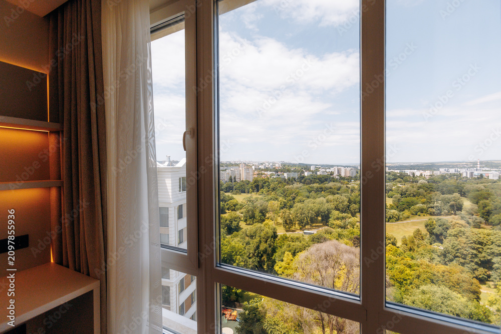 Fototapeta premium Hotel room window overlooking urban park, distant skyline, and clear blue sky with soft warm interior lighting, framed by sheer curtains, glass reflections, and spacious high floor view
