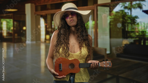 Fotografie Hispanic woman holding ukulele, wearing straw sun hat and sunglasses inside a hotel building lobby; vacation relaxed