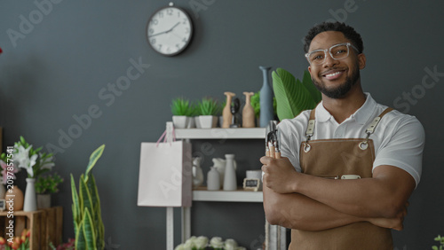Fotografie Man florist smiles with arms crossed holding pruning shears, bare forearms visible, wearing apron and glasses in a flower shop filled with potted plants and vases; welcoming warmth