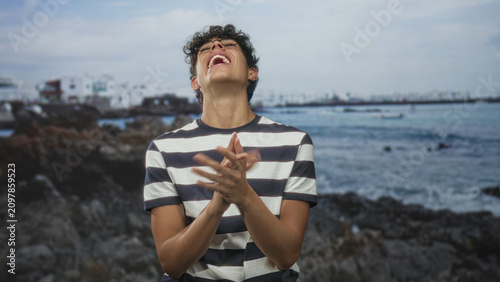 Konstfotografi Teenage boy laughing with hands on hips by seaside building, wearing striped shirt and glasses, facing camera; joy playfulness youth