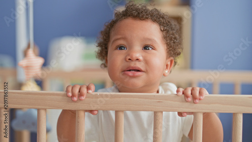 Canvas Print Little girl stands and grips crib rail with small hands in nursery building near mobile; tender curiosity warmth