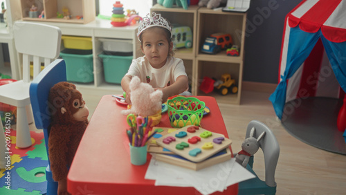 Canvastavla Hispanic girl in tiara plays with a teddy and colorful toy dishes at a red table in a bright playroom; pretend tea playful