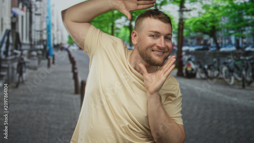 Quadro su tela Man framing face with hands and tilted head on cobbled street in beige tshirt smiling broadly with beard visible; playful confidence